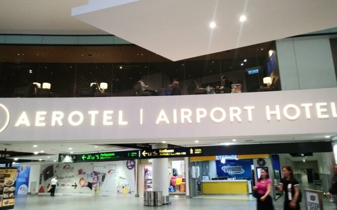 A large white and beige entrance sign to the Aerotel Kuala Lumpur Airport Hotel in the Gateway Mall with surrounding shops and people.