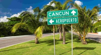 A green traffic sign pointing to the airport or Cancun with arrows on a grassy street corner.