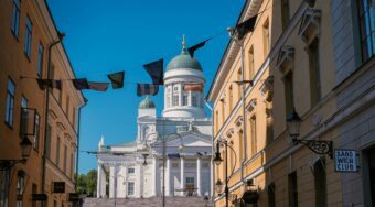 Fabric flags wave overhead in front of a white ornate building in Helsinki