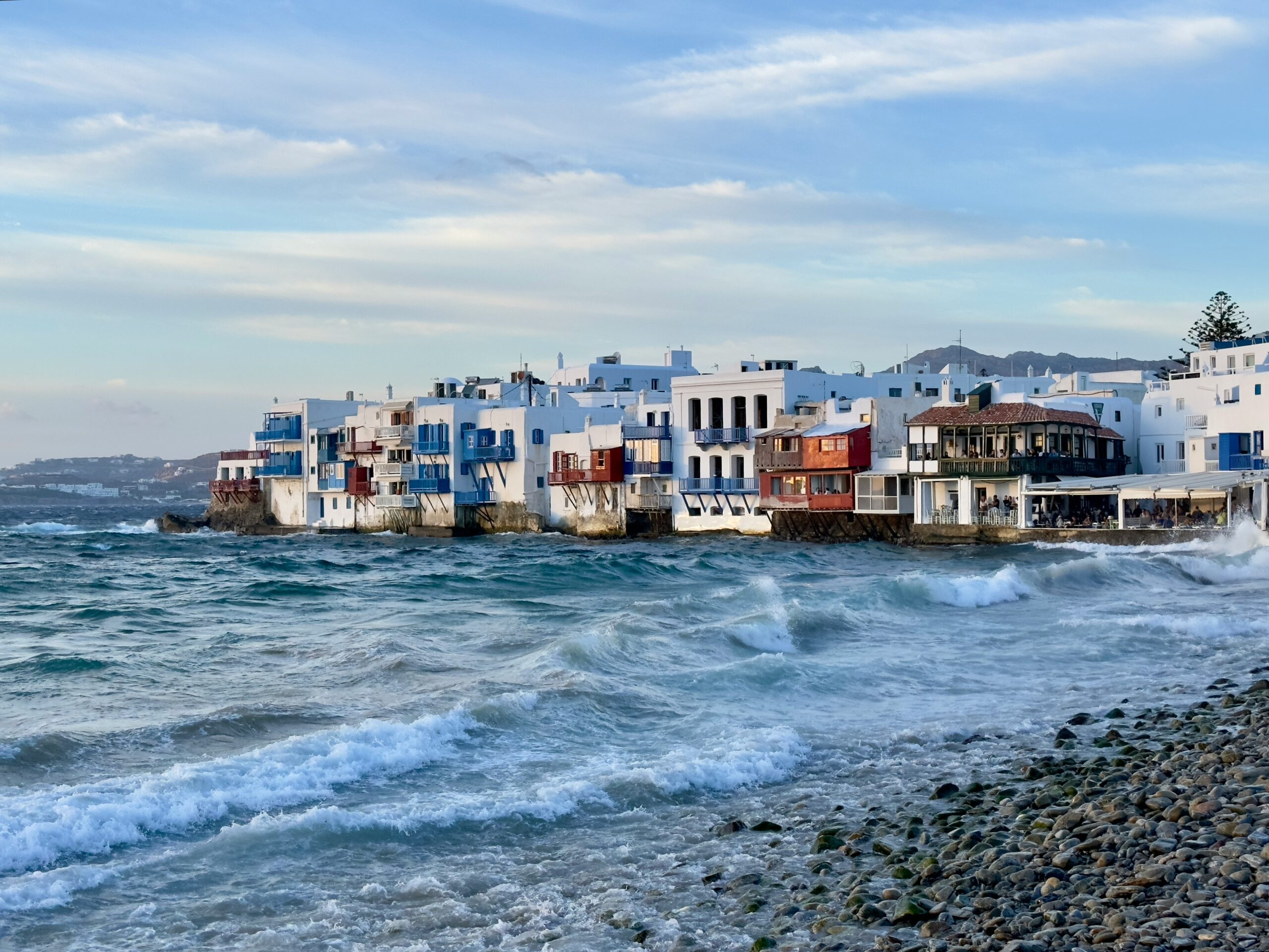 A pebbly beach with waves coming in and houses along the shore in the distance.