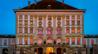 An ornate old building with lots of windows and a bridge in front lit up with pink lighting.
