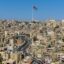 A view of Amman city filled with yellow, white and, beige buildings, and a Jordanian flag towering above the skyline.