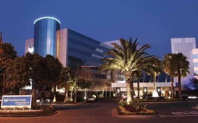 The front entrance to the Southern Sun Airport Hotel at Johannesburg Airport lit up at night with trees, palm trees and a sign in front.