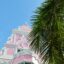 A pink and white ornate building against a blue sky with a palm tree in front.