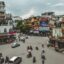 people riding on mopeds and vehicles at an intersection surrounded by colorful buildings in Hanoi