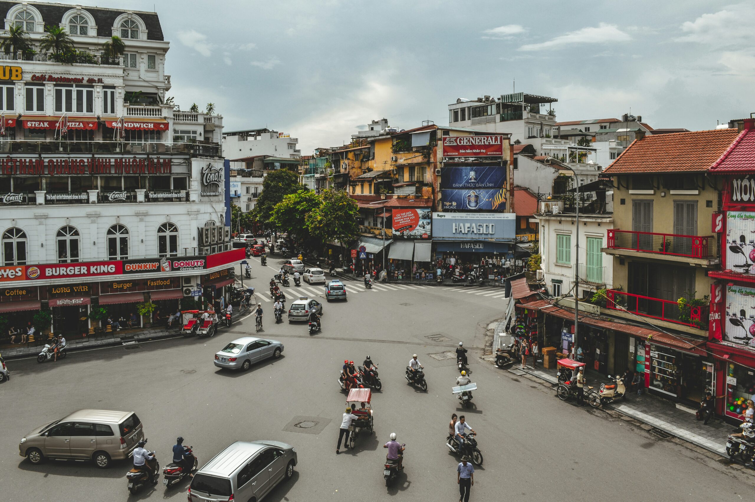 people riding on mopeds and vehicles at an intersection surrounded by colorful buildings in Hanoi
