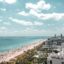 strip of light colored sand sandwiched by blue water and green trees scattered with buildings under a cloudy sky