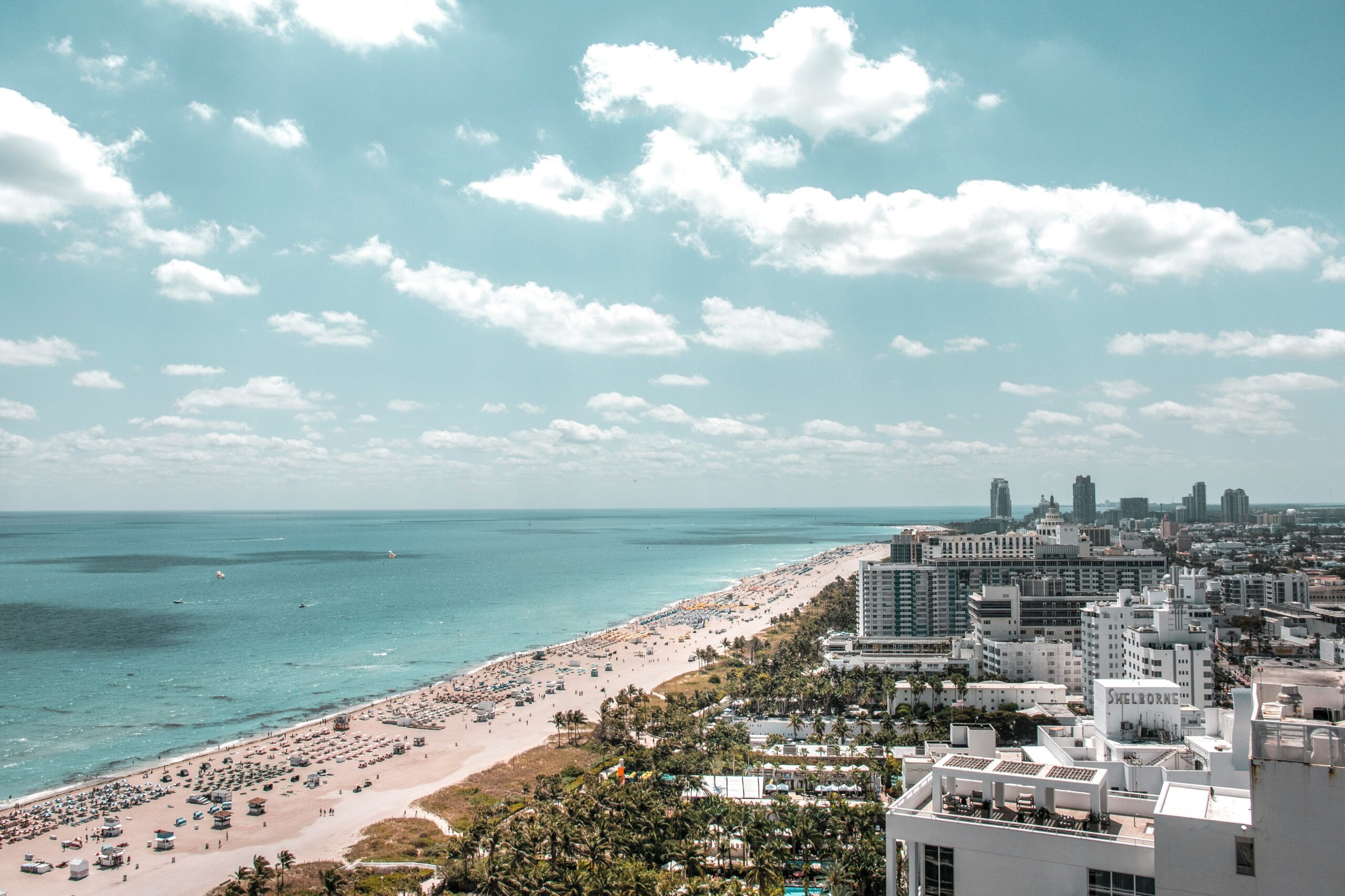 strip of light colored sand sandwiched by blue water and green trees scattered with buildings under a cloudy sky