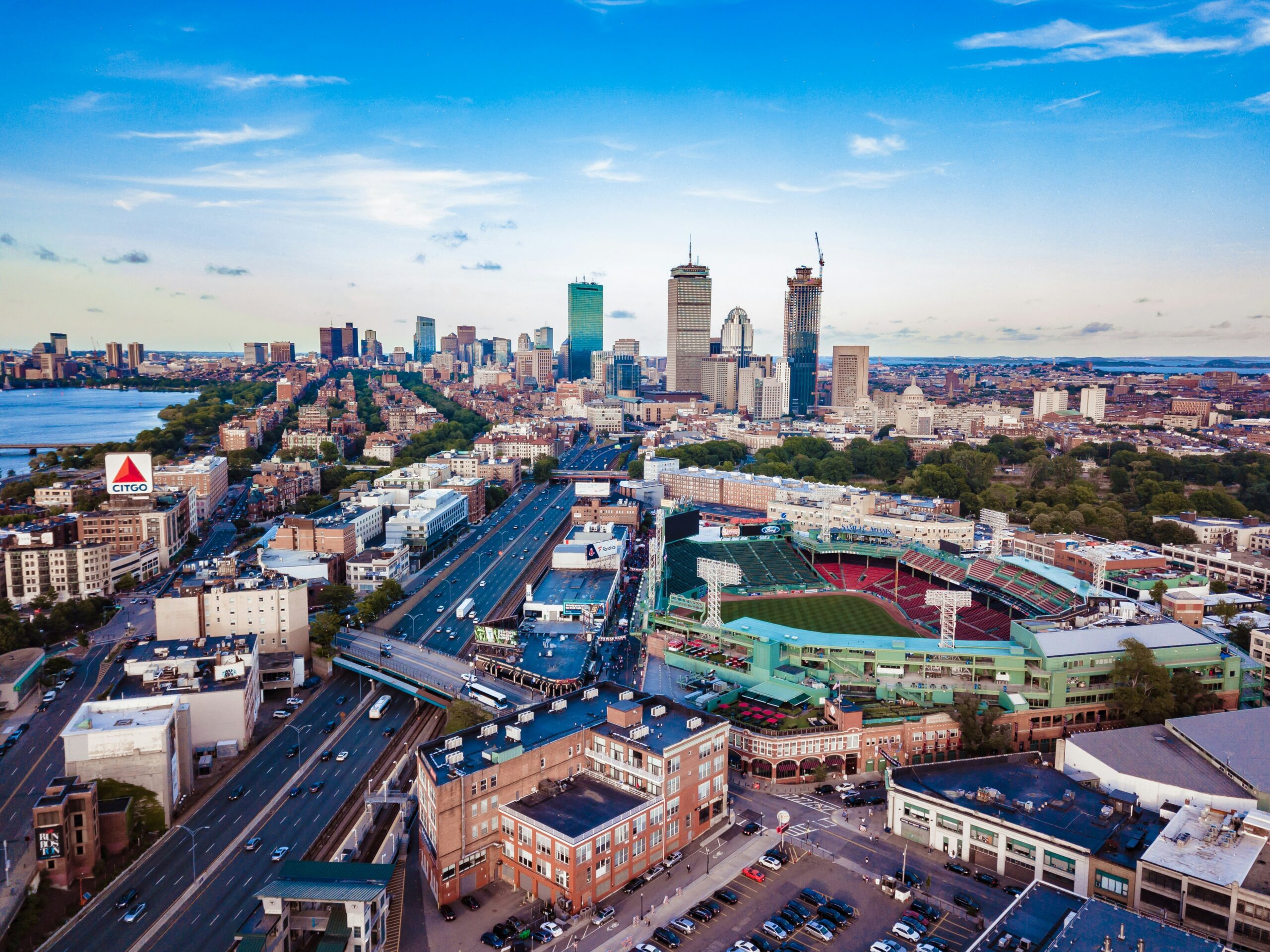 Aerial view of downtown Boston at sunset, featuring Fenway Park, the Charles River, and the Prudential Tower in the skyline. Major highways and city streets are visible, highlighting the accessibility and convenience of a Boston car service for navigating the urban landscape.