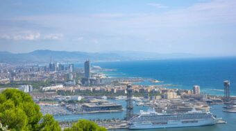 View of Barcelona Cruise Port from a hill, with a large cruise ship in the docks, buildings in the background, and a large body of water to the right.