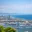View of Barcelona Cruise Port from a hill, with a large cruise ship in the docks, buildings in the background, and a large body of water to the right.