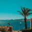 A view of the ocean with turquoise water framed by a blue sky, palm trees and square sandstone buildings.