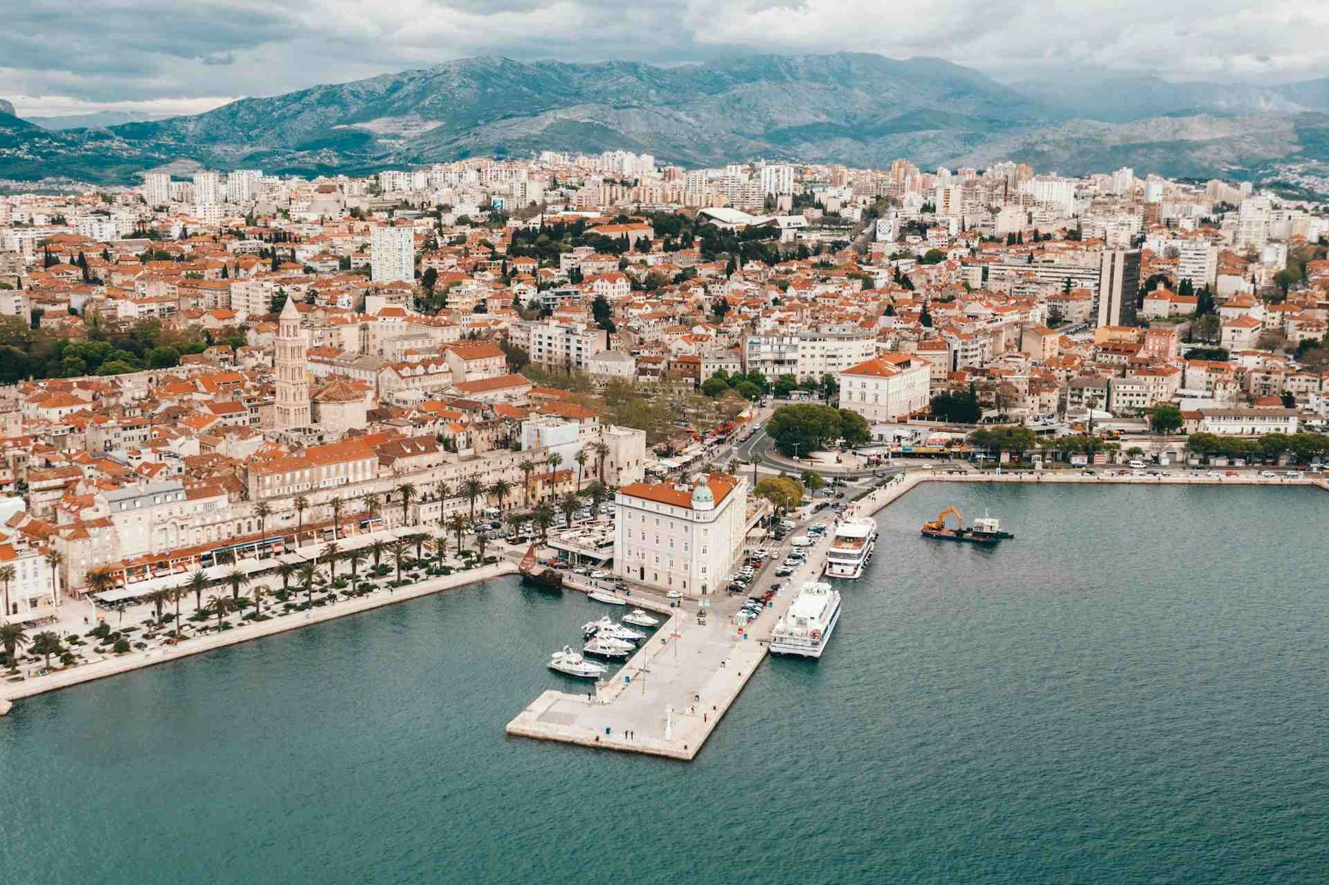 Aerial view of coastal Split, showing the port and buildings, with mountains in the background and water in the foreground.