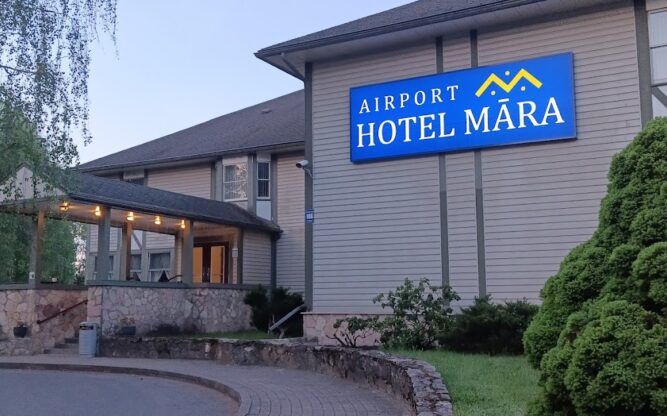 The entrance to the Airport Hotel Mara at Riga Airport. A brown building with a blue sign surrounded by trees.