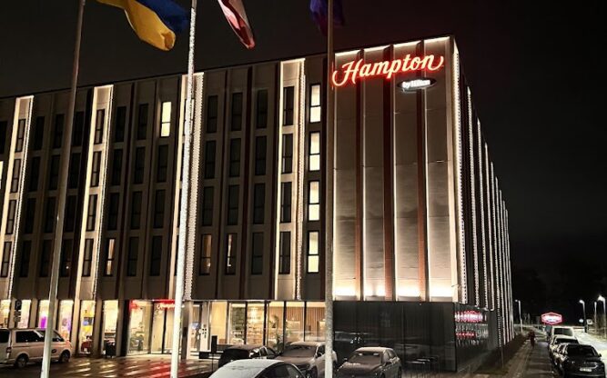 A tall rectangular building lit up at night with a lawn and two flag poles in the foreground