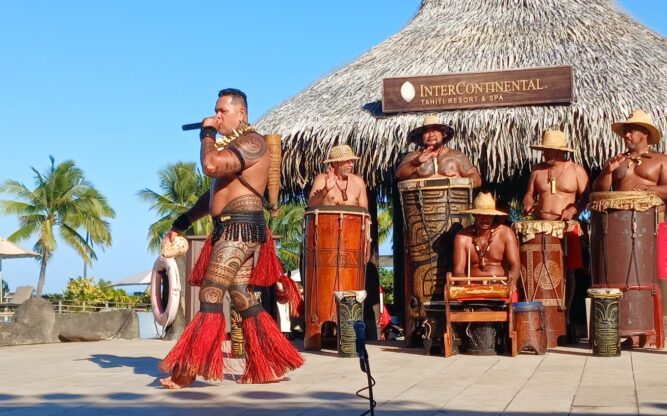 A group of men dressed in tribal clothing playing instruments in front of the Intercontinental Tahiti Airport Hotel.