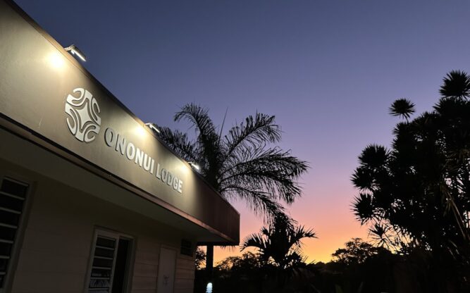 The front of the Ononui Lodge with a lit up a sign against the backdrop of a sunset sky and the silhouette of palm trees.