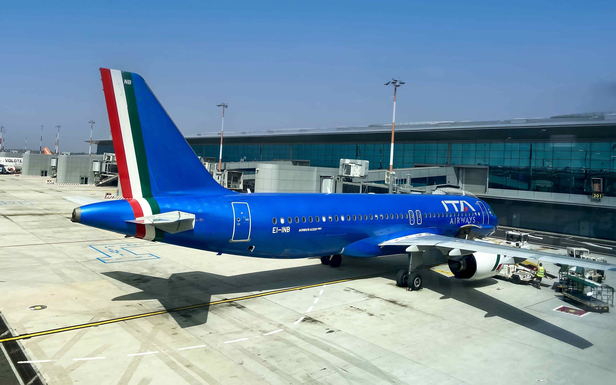 A blue ITA plane ready for departure, parked at a boarding gate, with blue skies above.