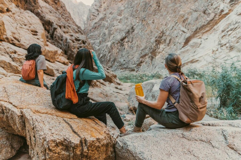 Three women on an expedition; wearing backpacks and holding water bottles while sitting on rocky terrain.
