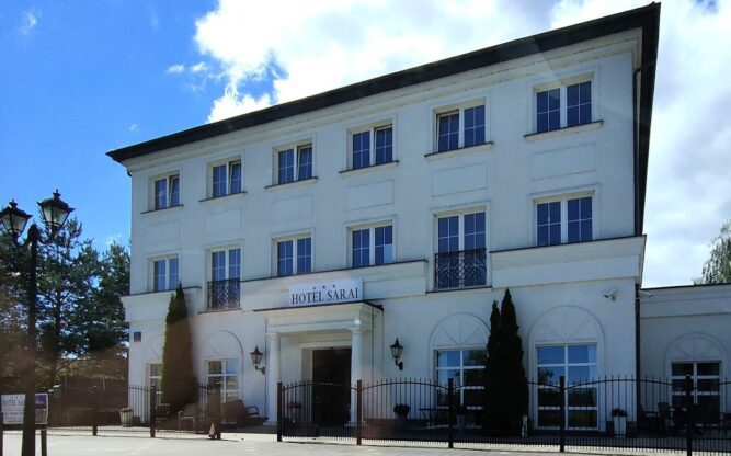 The charming white facade of the Hotel Sarai at Wroclaw Airport with lots of windows and a leafy driveway.