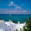 White square houses against the backdrop of the clear blue sea with a sail boat.