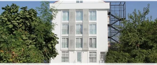 A white four-storey hotel with windows and small balconies with tall green bushes on either side