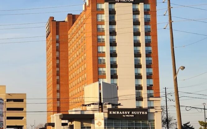 A tall beige and orange building on a street with power lines, an old car park and a few parked vehicles.