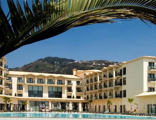 A large pool surrounded by a multi-storey cream and yellow resort and hotel with palm tree branch in front.