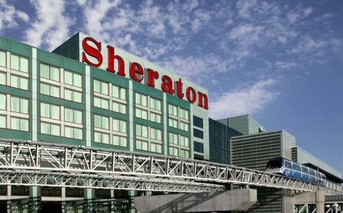 A large red "Sheraton" sign sits on top of a green building with lots of windows and a train running past in front.