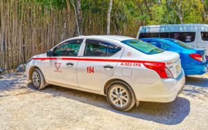 A white Cancun taxi parked on a gravel road near a bamboo fence, featuring a red stripe, official taxi markings, and license number—typical of Cancun taxi services used for local and airport transportation