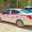 A white Cancun taxi parked on a gravel road near a bamboo fence, featuring a red stripe, official taxi markings, and license number—typical of Cancun taxi services used for local and airport transportation