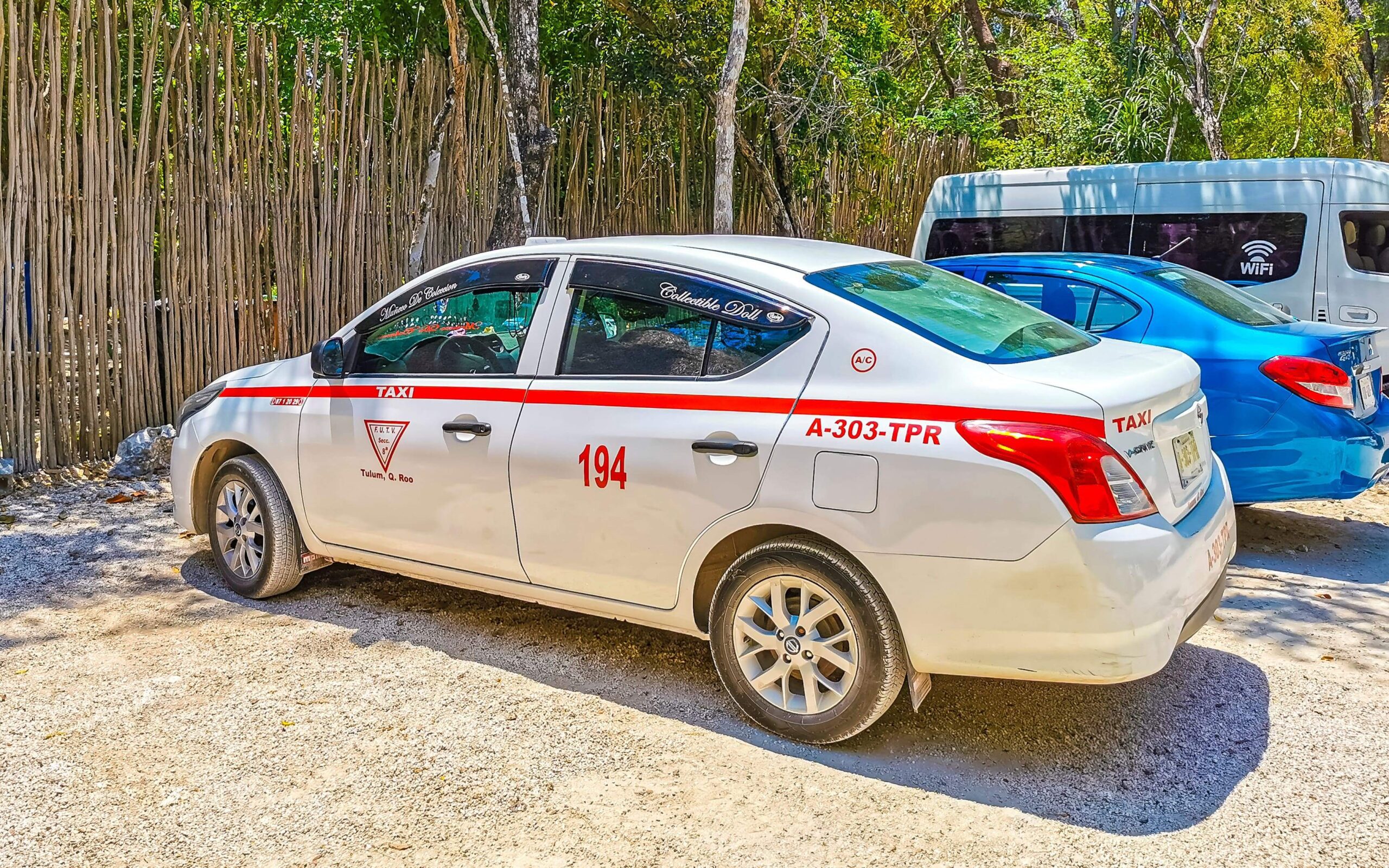 cancun taxi A white Cancun taxi parked on a gravel road near a bamboo fence, featuring a red stripe, official taxi markings, and license number—typical of Cancun taxi services used for local and airport transportation