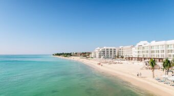 A panoramic view of the Cancun Hotel Zone showing modern beachfront resorts with white façades lining a pristine sandy beach and turquoise water under a clear blue sky — a popular destination for travelers making the journey from Cancun Airport to Hotel Zone.