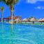 Scenic beachfront view at a luxury resort in Puerto Morelos, featuring clear turquoise water, palm trees, and thatched sunshades under a bright blue sky. A tranquil destination accessible via a 25-minute transfer from Cancun Airport to Puerto Morelos.