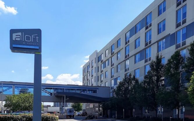 A driveway leading up to the Aloft Hotel with a blue sign and some lawn on the left.