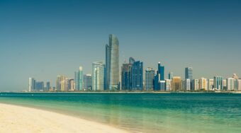 View of Abu Dhabi’s modern skyline with glass skyscrapers and clear blue waters seen from a sandy beach on a bright day, representing the final destination for travelers journeying from Dubai International Airport to Abu Dhabi.