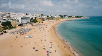 Wide-angle view of Albufeira’s golden sandy beach on a bright afternoon, with sunbathers, colorful umbrellas, and clear turquoise water stretching along the coast. Whitewashed buildings and cliffside hotels overlook the shoreline, capturing the relaxed atmosphere typical of a summer arrival from Faro Airport to Albufeira.