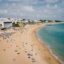 Wide-angle view of Albufeira’s golden sandy beach on a bright afternoon, with sunbathers, colorful umbrellas, and clear turquoise water stretching along the coast. Whitewashed buildings and cliffside hotels overlook the shoreline, capturing the relaxed atmosphere typical of a summer arrival from Faro Airport to Albufeira.
