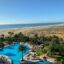 A sunny daytime view of a beachfront resort in Costa Calma, Fuerteventura, featuring a large pool surrounded by palm trees and sun loungers, with golden sands and the Atlantic Ocean in the background. This peaceful coastal retreat is located about an hour's drive south of Fuerteventura Airport.