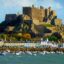 View of Mont Orgueil Castle overlooking the harbor and colorful seafront houses in Gorey, Jersey, with boats anchored in the bay on a clear afternoon. A popular destination easily reached by Jersey Airport taxi for travelers exploring the island’s historic sites.