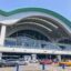 Exterior view of Istanbul Sabiha Gokcen Airport during the day, showing the curved roof design, glass façade, and busy passenger drop-off area with taxis and cars.