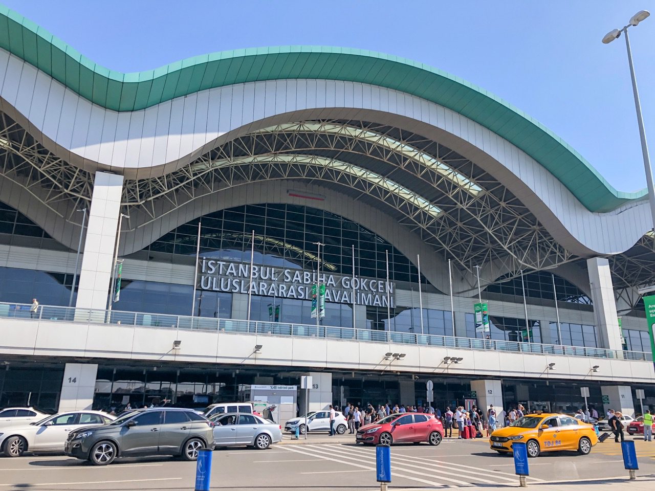 Exterior view of Istanbul Sabiha Gokcen Airport during the day, showing the curved roof design, glass façade, and busy passenger drop-off area with taxis and cars.