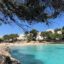 Sunny afternoon at a beach in Cala d'Or, Mallorca, with turquoise waters, rocky cliffs, and white resort buildings surrounded by pine trees—showcasing the coastal charm awaiting travelers journeying from Mallorca Airport to Cala d'Or.