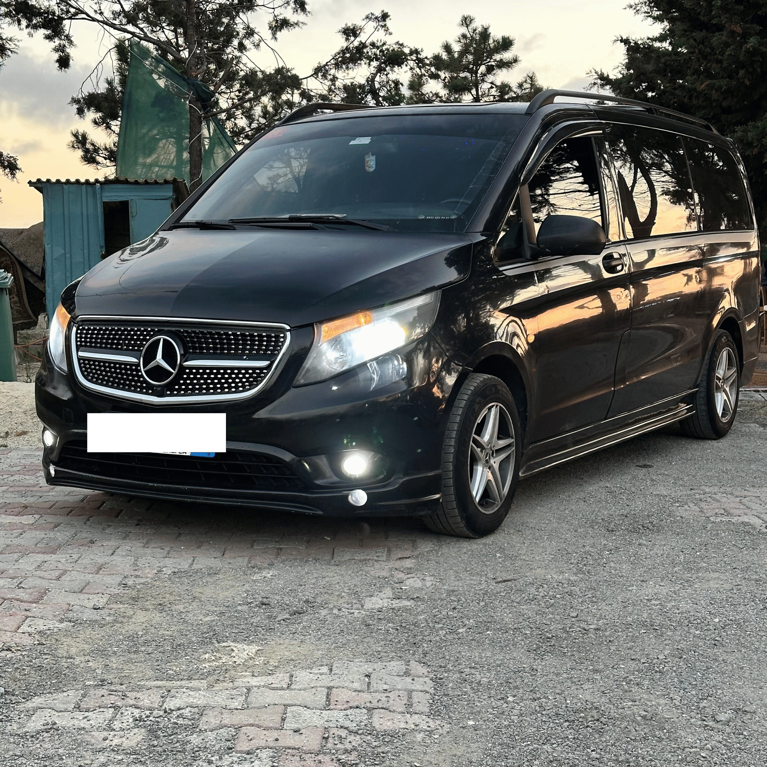 A black Mercedes van with headlights on, parked on a quiet roadside at sunset—commonly used as an airport taxi in Istanbul for convenient city and airport rides.
