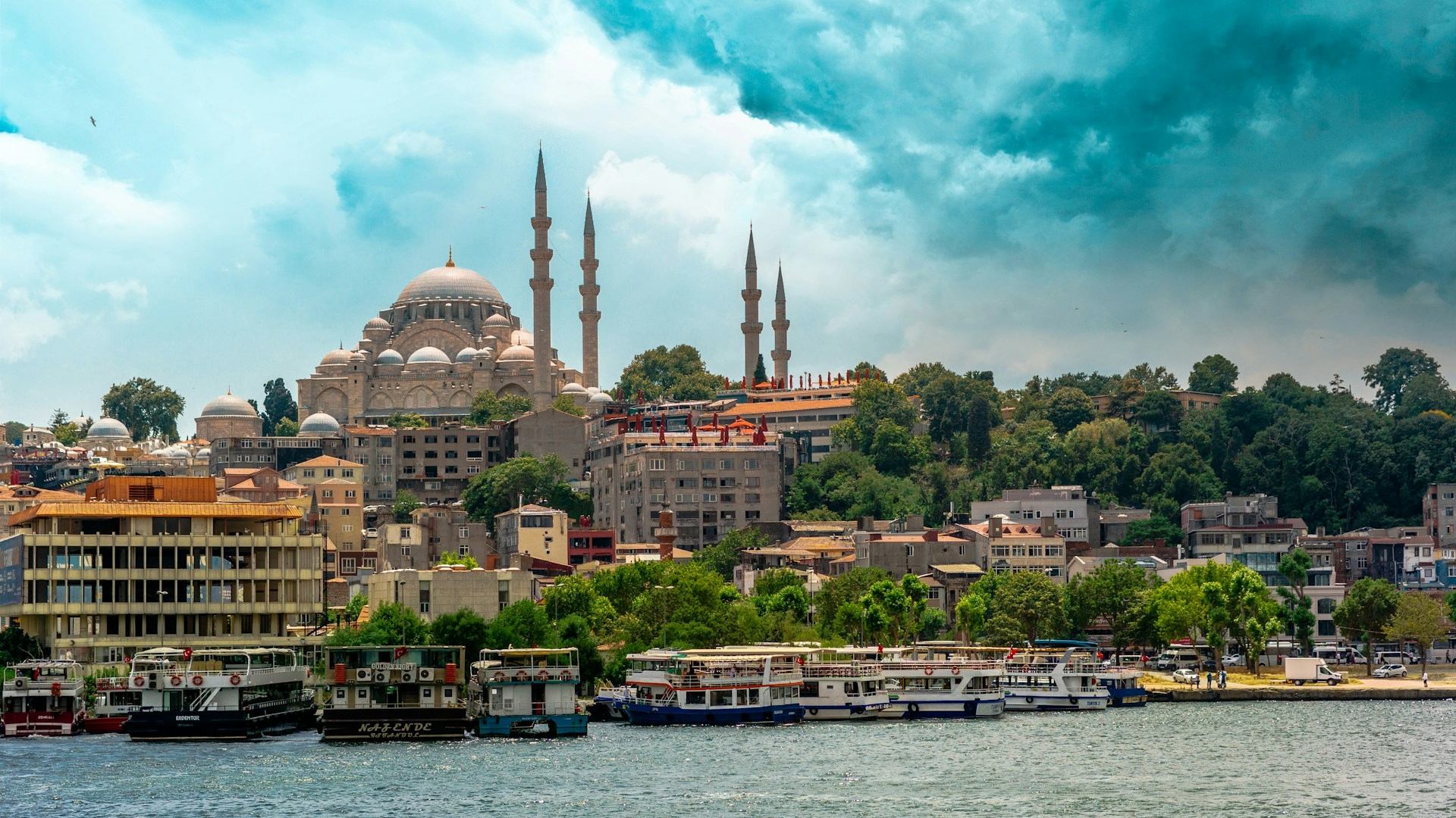 Scenic daytime view of Istanbul’s historic Süleymaniye Mosque rising above a lively mix of waterfront buildings, trees, and docked boats along the Golden Horn — a striking first impression for travelers enjoying an airport transfer in Istanbul.