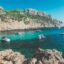 Scenic coastal view near Mallorca featuring turquoise waters, rocky cliffs, and several small boats anchored in a calm bay under a clear blue sky; a popular destination easily reached with a Mallorca Airport transfer.