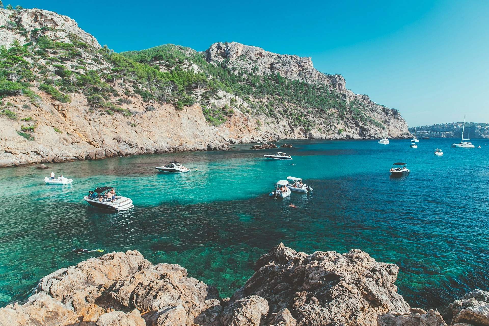 Scenic coastal view near Mallorca featuring turquoise waters, rocky cliffs, and several small boats anchored in a calm bay under a clear blue sky; a popular destination easily reached with a Mallorca Airport transfer.
