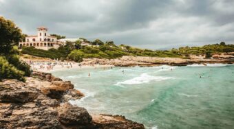 View of a rocky coastline and sandy beach near Alcudia on an overcast day, with turquoise waves, swimmers in the sea, and a Mediterranean-style villa overlooking the shore.