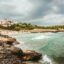 View of a rocky coastline and sandy beach near Alcudia on an overcast day, with turquoise waves, swimmers in the sea, and a Mediterranean-style villa overlooking the shore.