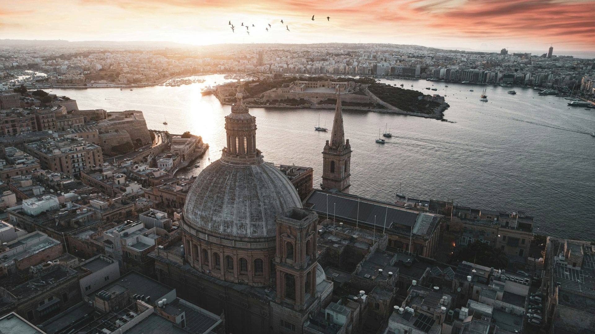 A panoramic view of Valletta, Malta at sunset, with historic domes, church spires, and the harbor in the foreground. Travelers booking a Malta airport transfer can enjoy this scenic arrival to the city, with boats on the water and a warm evening sky overhead.